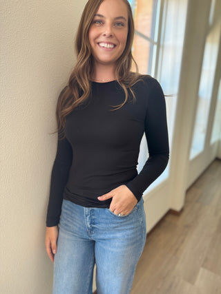 Woman wearing a black long-sleeve top and blue jeans standing against a neutral wall.