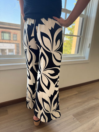 Person wearing black and white floral pants standing in a room with a window in the background.