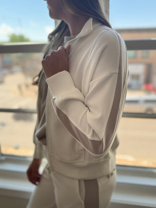 Person wearing a beige tracksuit standing by a window with a blurred outdoor view.