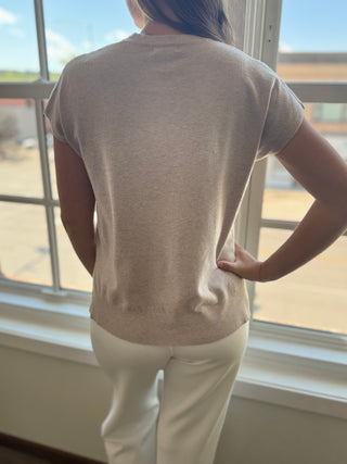 Person wearing a beige sweater and white pants standing by a window.