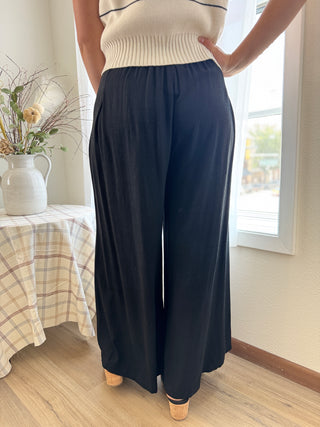 Person wearing navy wide-leg pants and a cream top in a room with a window and table.