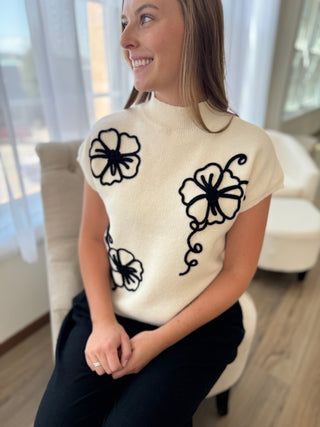 Woman wearing a cream sweater with black floral patterns in a room with large windows.