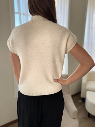 Person wearing a beige short-sleeve sweater in a room with white curtains.