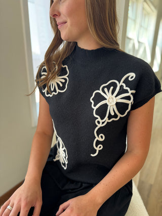 Woman wearing a black top with white floral embroidery sitting indoors.