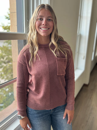 Woman wearing a pink sweater and blue jeans standing indoors near a window.