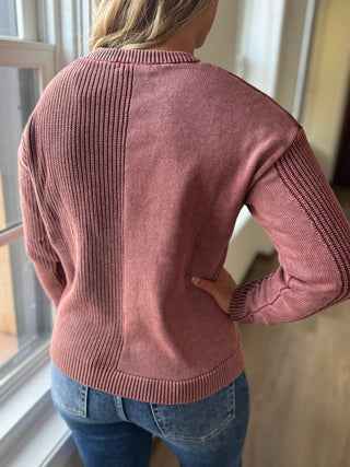 Person wearing a pink sweater and blue jeans standing by a window.