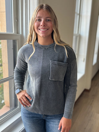 Woman wearing a gray sweater with a pocket, standing indoors by a window.