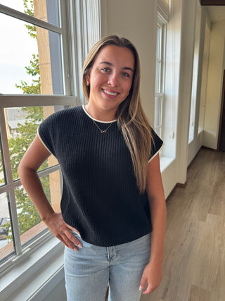 Woman standing in a room with large windows, wearing a dark ribbed sweater and light jeans.
