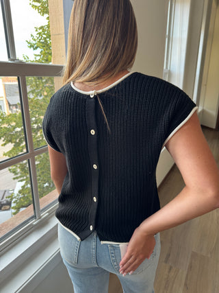 Person wearing a black ribbed sweater with white buttons and light blue jeans indoors.