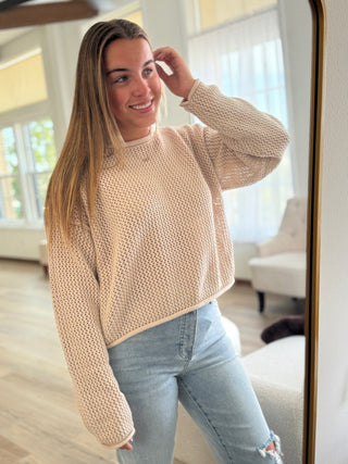 Woman wearing a beige sweater and light blue jeans standing in a room with a mirror.