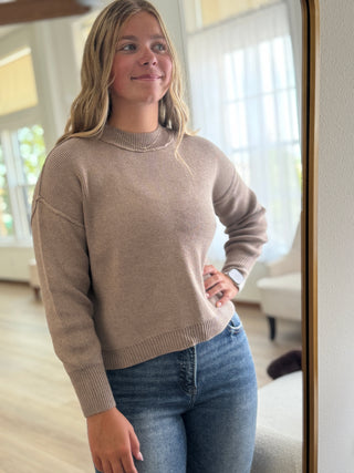 Woman wearing a beige sweater and blue jeans standing in front of a mirror.