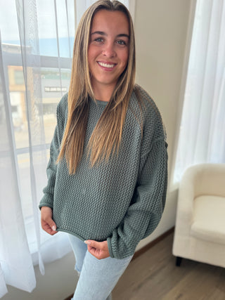 Woman wearing a green sweater in a room with white curtains and a beige chair.