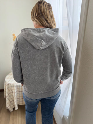 Person wearing a gray hoodie and blue jeans standing in a room with a curtain.