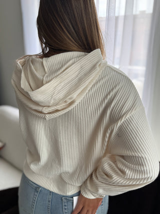 Person wearing a beige ribbed hoodie and blue jeans in a softly lit room.