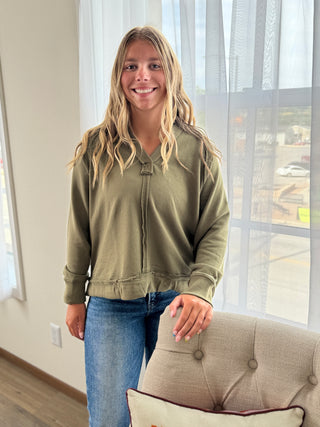 Woman wearing a green jacket and blue jeans standing in a room with a couch and window.
