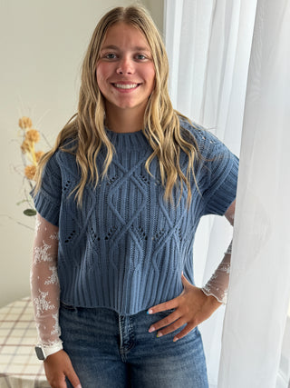 Woman wearing a blue knitted top with a white lace sleeveless top underneath, standing indoors.