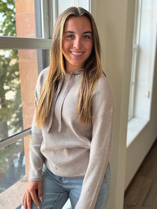 Woman wearing a beige long-sleeve top and jeans standing by a window.
