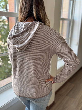 Person wearing a beige hoodie standing in a room with large windows.