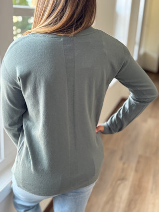 Person wearing a green long-sleeve shirt in a room with wooden flooring and a window.