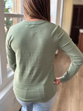 Person wearing a green long-sleeve shirt standing indoors near a window.
