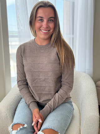Woman sitting on a chair wearing a brown sweater and ripped jeans, smiling at the camera.