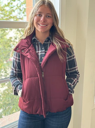 Person wearing a maroon puffer vest over a plaid shirt indoors.