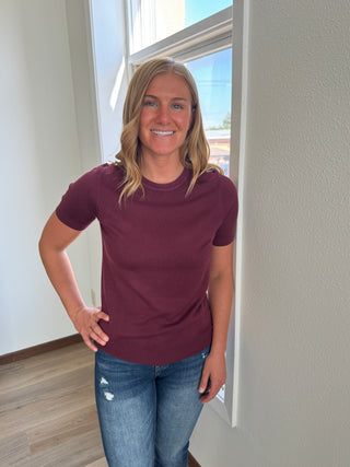 Woman wearing a maroon shirt and blue jeans standing in a room with a window.