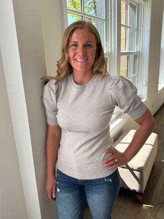 Woman in a gray sweater and blue jeans standing indoors with a neutral background