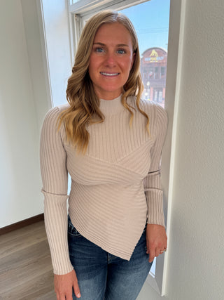 Woman wearing a beige ribbed sweater and blue jeans standing in a room with a window.