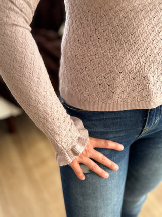 Person wearing a beige cable knit sweater and blue jeans with a blurred background