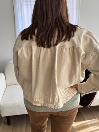 Person wearing a beige jacket and brown pants sitting on a white couch.