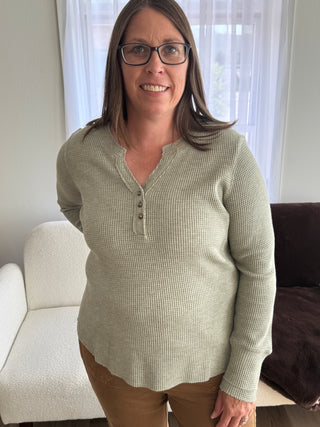 Woman wearing a beige sweater standing indoors with a window in the background