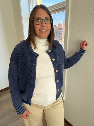 Woman wearing a navy cardigan over a white sweater, standing indoors with a window in the background.