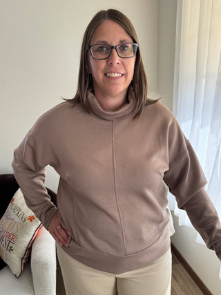 Woman wearing a beige sweater and glasses, standing indoors with a neutral background