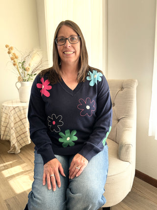 Woman wearing a navy sweater with colorful flower patterns, sitting on a couch in a living room.