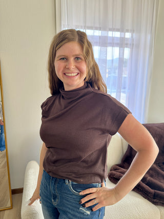 Woman wearing a brown top and blue jeans standing in a room with a window in the background.