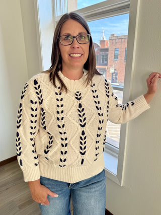 Woman wearing a cream sweater with black patterns standing indoors near a window.