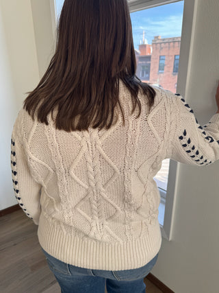 Person wearing a beige cable knit sweater with a blurred background