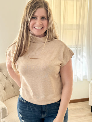 Woman wearing a beige turtleneck top and blue jeans indoors.