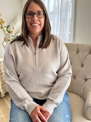 Woman wearing a beige pullover with a zipper, sitting on a couch in a home setting.