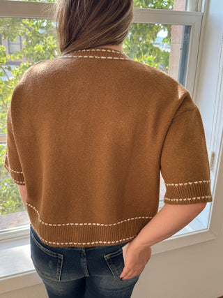 Person wearing a brown sweater with white trim, standing indoors near a window.