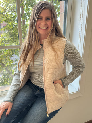 Woman wearing a beige quilted vest and gray sweater indoors with a window in the background.