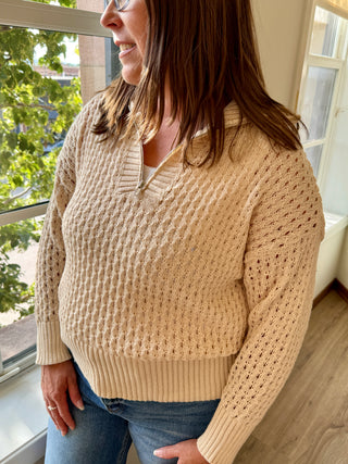 Woman wearing a beige knitted sweater standing by a window with a view of trees outside.