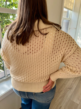 Person wearing a beige knitted sweater and blue jeans standing by a window.