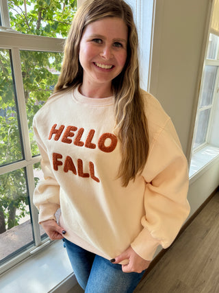 Person wearing a 'Hello Fall' sweatshirt standing by a window with greenery outside.