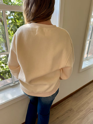 Person wearing a beige sweater and blue jeans standing in front of a window with a view of trees.
