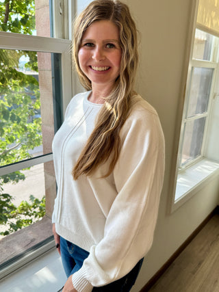 Woman in a white sweater standing by a window with a view of trees