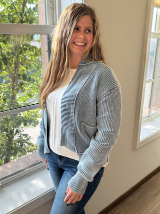 Woman wearing a gray sweater and blue jeans standing indoors with large windows.