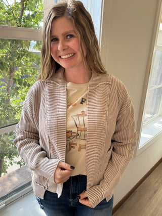 Woman wearing a beige cardigan over a graphic t-shirt indoors with a window in the background