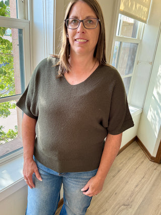 Woman wearing a brown sweater and blue jeans standing in a room with large windows.